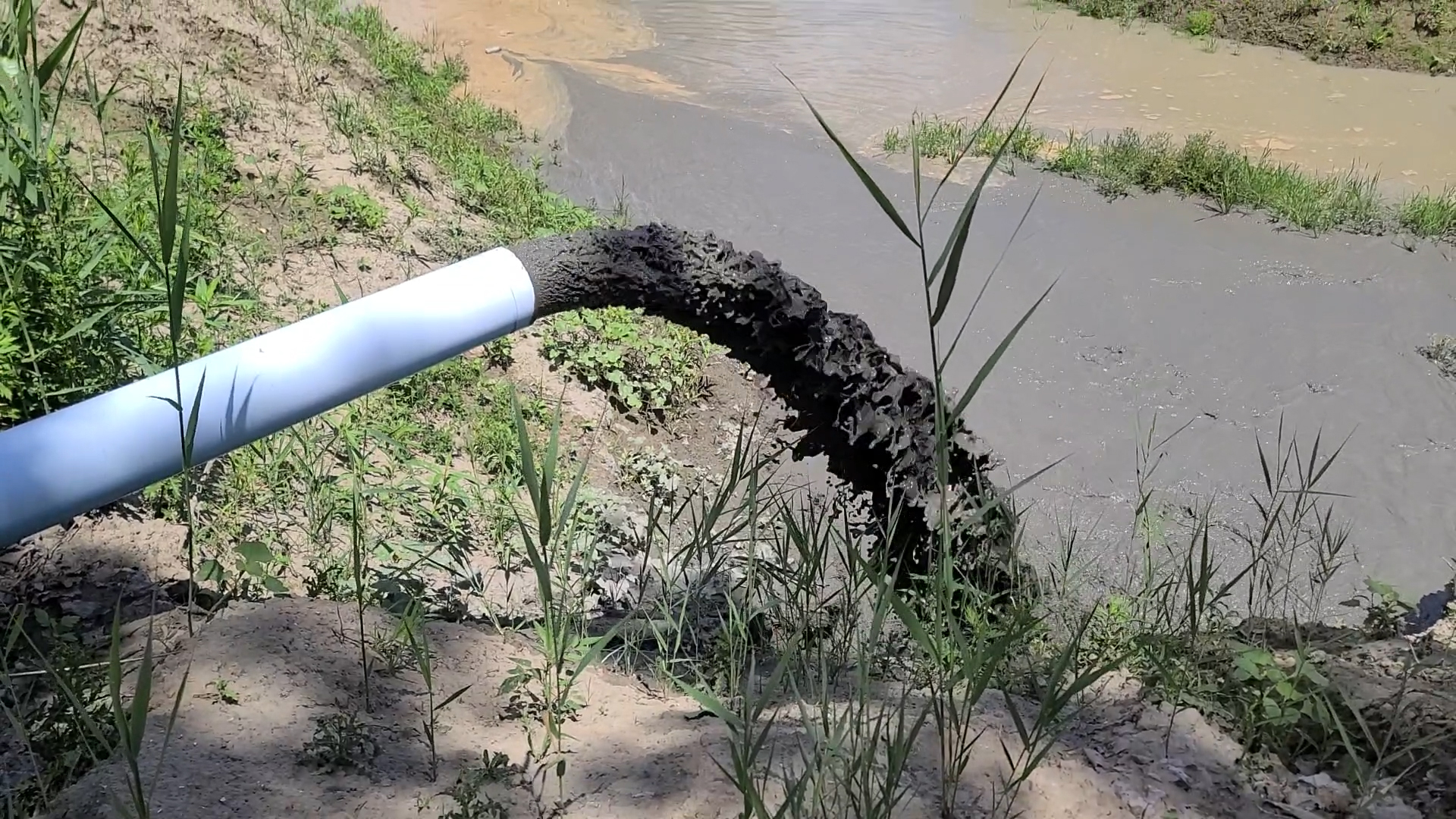 "Pipe Discharging Dark Slurry into Muddy Water with Vegetation Nearby " A pipe discharging dark slurry into a muddy water body