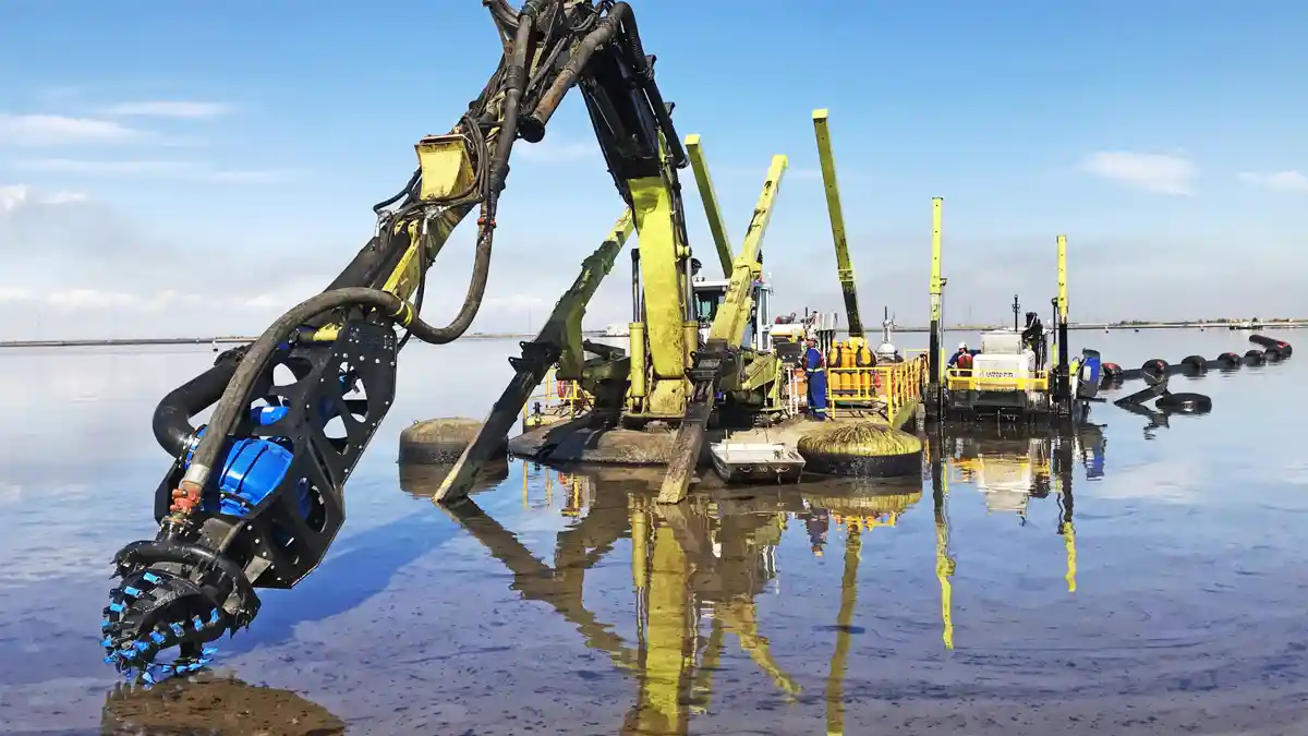 Amphibious excavator on spud legs raising an EDDY Pump dredge attachment and cutterhead from shallow water